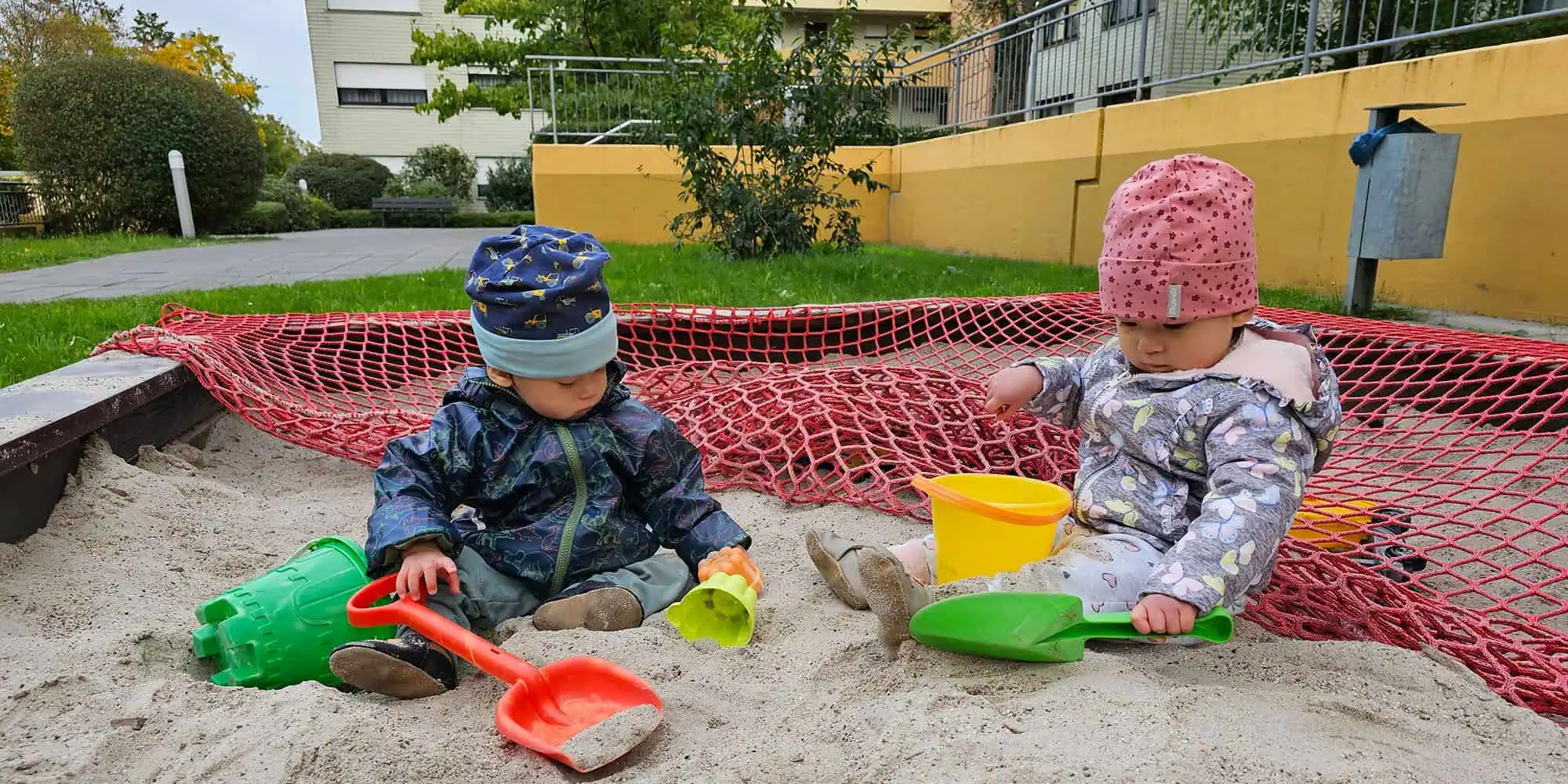 Zwillingsbabys im Sandkasten, eines in Rosa und eines in Blau gekleidet.