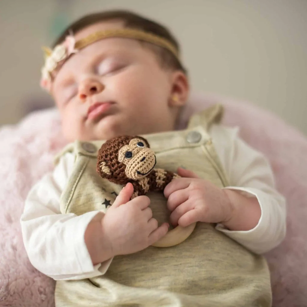 Schlafendes Baby hält die Ringrassel „Affe Charlie“ mit beiden Händen; die Rassel ist scharf, das Baby weich im Hintergrund.