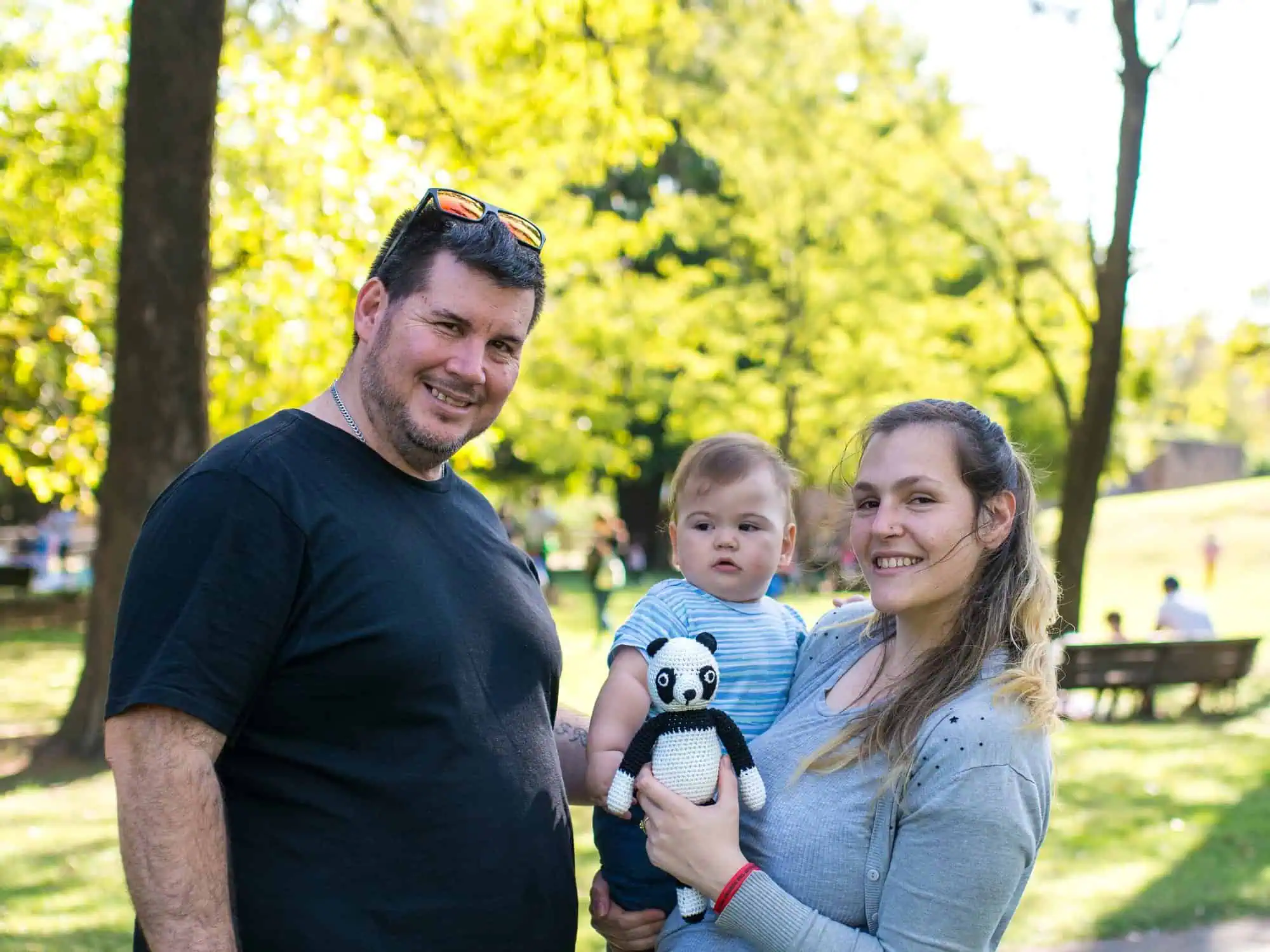 Happy family in the park: mother holding a SindiBaba panda cuddle toy, baby in her arms, father standing beside them.
