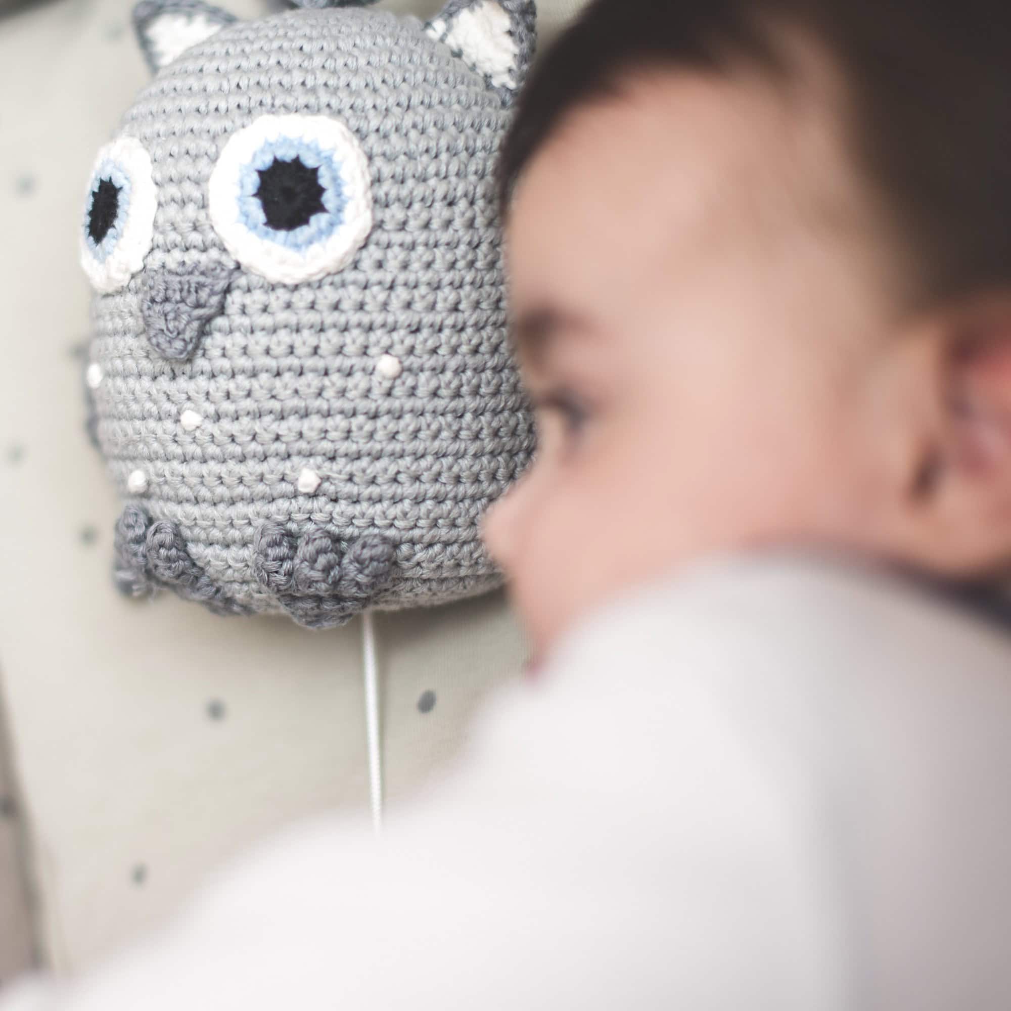 Sleepy toddler lying in bed next to the grey SindiBaba musical toy “Owl”; owl in focus, child softly blurred in the foreground.