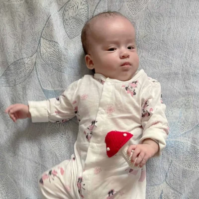 12425-baby-rattle-toadstool-lifestyle-baby-girl Babymädchen liegt auf gemusterter Decke und hält eine gehäkelte Fliegenpilz-Babyrassel