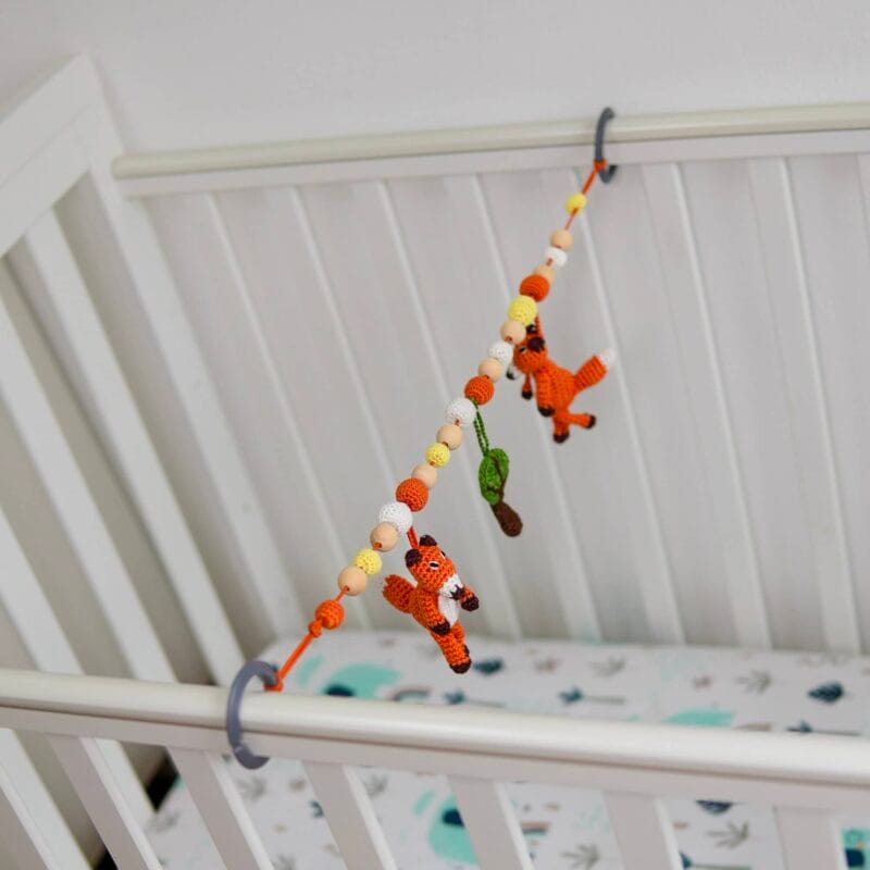 12546-stroller-chain-fox-fred-lifestyle-baby-cot Sehr helle Lifestyle-Aufnahme der elastischen Fuchs FRED Kinderwagenkette, die mittels grauen Ringen über die volle Länge eines weißen Babybettes gespannt ist.