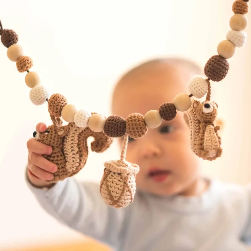 12559-stroller-chain-squirrel-nutty-lifestyle-playgro-eye-level Lichtdurchflutetes Premium-Lifestyle-Foto auf Augenhöhe: Ein Kleinkind sitzt auf hellem Holzfußboden und greift nach der Eichhörnchen Kinderwagenkette vor fast weißem Hintergrund.
