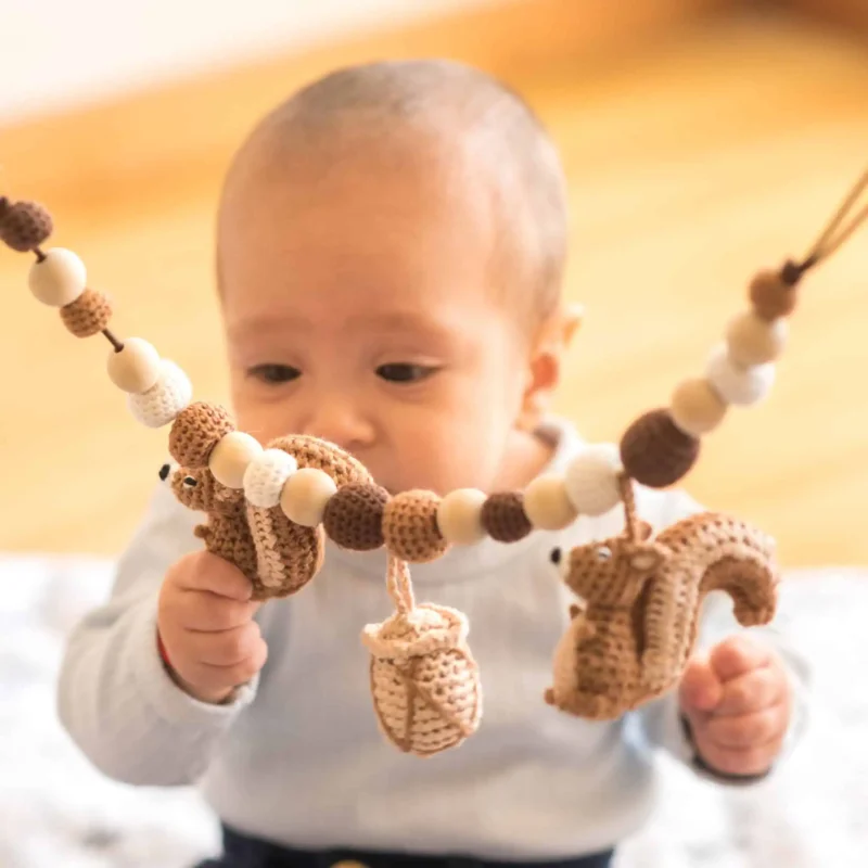 12559-stroller-chain-squirrel-nutty-lifestyle-playgro-high-angle DE: Helles Lifestyle-Foto aus erhöhter Kameraperspektive: Das Kleinkind sitzt auf einer hellen Decke auf dem honigfarbenen Holzfußboden und spielt mit beiden Händen an der Kette.