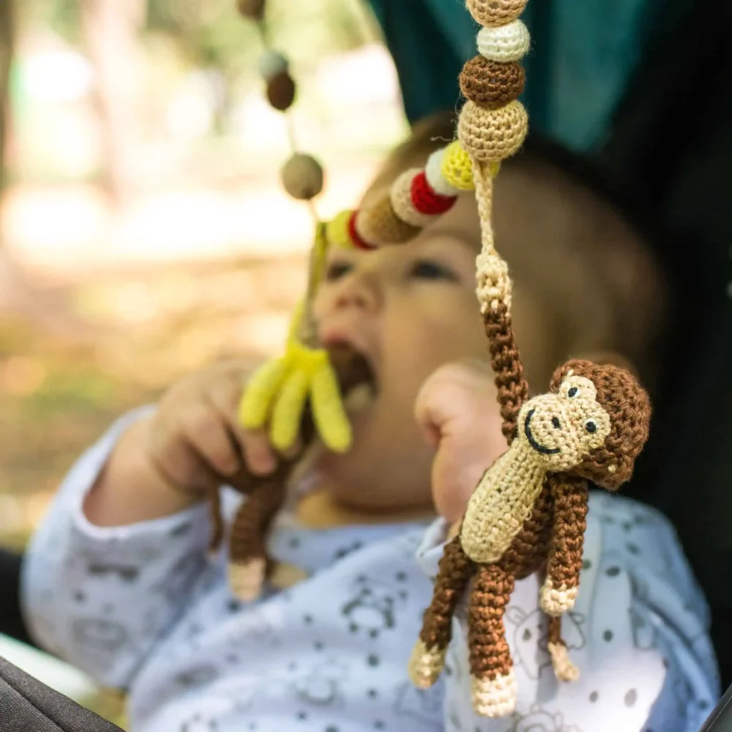Baby sitting sideways in the stroller and chewing on the monkey of the SindiBaba stroller chain; park with trees and shade softly blurred in the background.